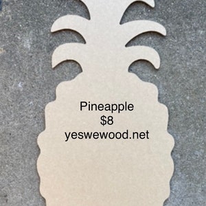 May include: A light brown wooden pineapple cutout. The cutout has a textured surface and the text "Pineapple" is printed on the front. The website yeswewood.net is also printed on the front.
