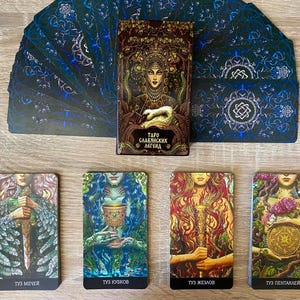 May include: A tarot card deck with a brown box featuring a woman with a crown and the text "TARO СЛАВЯНСКИХ ЛЕГЕНД". The deck includes cards with illustrations of figures and symbols, with a blue and silver design on the back.