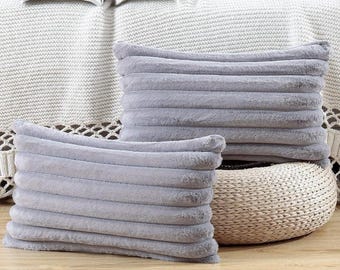 May include: Two gray, rectangular throw pillows with a ribbed texture. The pillows are made of a soft, plush material. One pillow is in front of the other. The pillows are on a wooden surface.