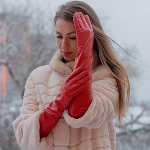 Può includere: Un paio di lunghi guanti in pelle rossa liscia che arrivano oltre i gomiti, indossati con un cappotto in pelliccia sintetica testurizzata color beige chiaro con collo alto. I guanti presentano una silhouette aderente e una finitura lucida.