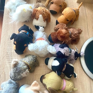 May include: A collection of fifteen small plush dog toys in various colors including tan, brown, black, white, and gray. Each plush toy features a soft texture, distinct facial features, and a small collar around the neck, arranged in a group.