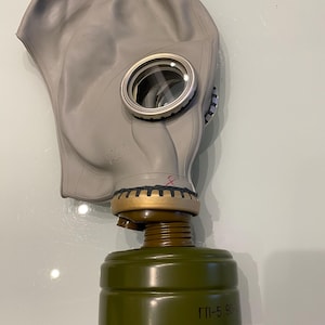 Op de afbeelding: Een grijze gasmasker met heldere ooglenzen en een groene filterbus. Het masker heeft een rubberen gezichtsbedekking en een metalen rand rond de lenzen. De filterbus heeft de tekst "ГП-5 90" erop.