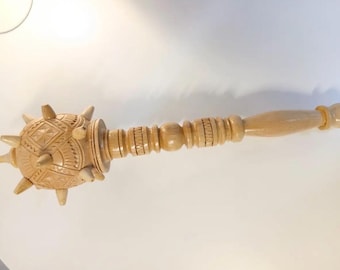 May include: A light-colored wooden mace with a spiked ball head and a long, carved handle. The ball head features intricate geometric carvings and is adorned with several pointed spikes. The handle has detailed carved rings.