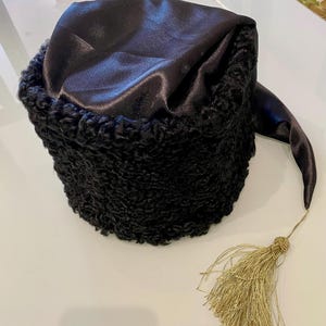 Puede incluir: Un sombrero de piel negro con una parte superior de satén negro y una borla dorada. El sombrero tiene una cola larga que se puede atar debajo de la barbilla.