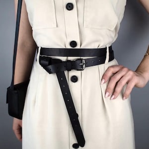 May include: A black leather belt with a silver buckle is wrapped around a cream dress. The belt's long end hangs down, and the dress has black buttons. The belt is approximately 2.5 cm wide.