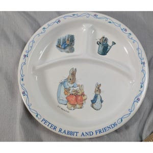 May include: White divided plate featuring illustrations of Peter Rabbit and friends. The plate has a blue floral border and the text "Peter Rabbit and Friends". The plate has three sections for food.