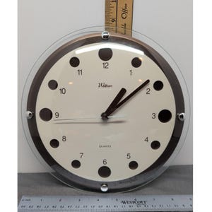 May include: A round Waltham quartz wall clock with a cream-colored face, dark brown hour markers, and black hands. The clock features a clear glass outer layer secured by four silver-tone metal studs. A ruler shows the clock is 10 inches in diameter.