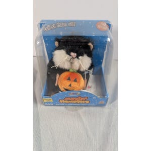 May include: A black and white plush hamster toy holding an orange pumpkin. The toy is in a clear plastic case with the text "Collect them all!" and "Mazinz Hamsters". The pumpkin has carved triangle eyes and a mouth.