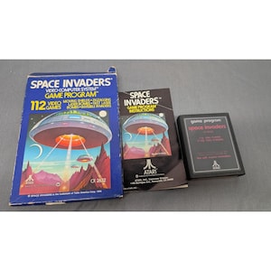 Puede incluir: Un set de videojuego Atari Space Invaders que incluye una caja de cartón azul, un manual de instrucciones y un cartucho de juego negro. La caja y el manual muestran un gráfico de un platillo volador con el texto Space Invaders.