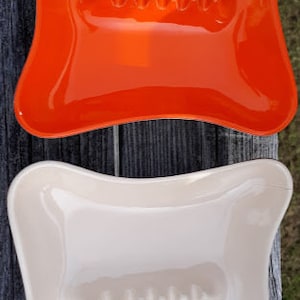 May include: Two ceramic ashtrays, one orange and one white, with a wavy edge and a raised center section. Both ashtrays have a raised, textured design in the center.