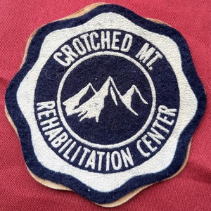 May include: A white felt patch with a blue circle in the center. The circle has a white mountain range design and the text "Crotched Mt. Rehabilitation Center" around the outside.