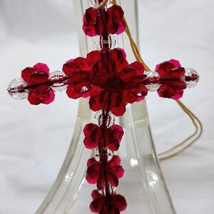 May include: A handcrafted cross pendant made of faceted red and clear beads. The cross features a floral design at the center and ends of each arm. A tan cord is attached for hanging. The pendant is displayed on a clear stand.