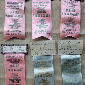 May include: A collection of vintage delegate ribbons from the Maine State Grange. The ribbons are pink and light blue, with text in green and gold. Each ribbon has the text "DELEGATE" and "MAINE STATE GRANGE". The ribbons also have the text "AUGUSTA DEC. 1-2-3, 1942" and "DEC. 7-9, 1943".