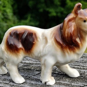 May include: A ceramic figurine of a collie dog standing in profile. The dog has a white base coat with patches of brown and dark brown fur on its back, neck, and head. The figurine features a glossy finish and is sculpted with detailed fur texture.
