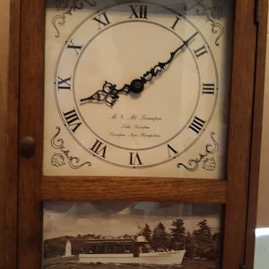 May include: A wooden clock with a white face and black Roman numeral hour markers. The clock has a black hand and a decorative design around the edge. The clock is mounted on a wooden frame with a picture of a boat on the water below it. The picture has the text "M.Y. "Mt. Tammany" Lake Champlain Champlain, New York"