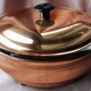 May include: A copper serving dish with a glass lid and black handles. The dish has a shiny, polished finish and is decorated with horizontal lines.