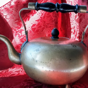May include: A vintage brass teapot with a wooden handle and a black knob on the lid. The teapot is sitting on a red fabric background.