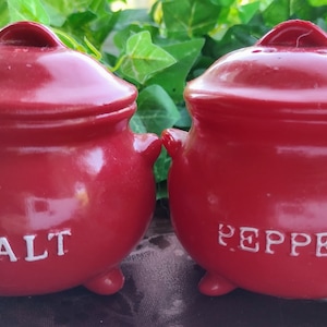 May include: Red ceramic salt and pepper shakers shaped like small pots with white lettering that reads "SALT" and "PEPPER".