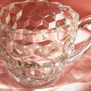 May include: A clear glass creamer with a diamond pattern and a handle. The creamer is sitting on a pink surface.