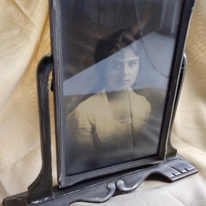 May include: A vintage black picture frame with a stand, featuring a black and white photo of a woman. The glass in the frame is cracked.