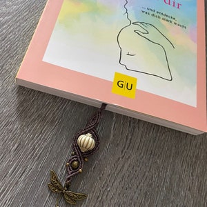 Peut inclure: Un livre rose avec le titre "Das Kind in dir... und entdecke, was dich stark macht" et les lettres "G/U" sur la couverture. Un marque-page en perles brunes et blanches avec un charme de libellule est attaché au livre.
