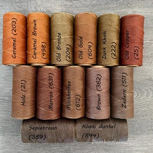 May include: A collection of fifteen cylindrical spools of thread in various shades of brown, tan, and copper. Each spool is labeled with a color name and a numerical code, including Caramel (203), Caramel Brown (498), Old Bronze (204), Old Gold (604), Dark Khaki (222), Old Copper (25), Holz (21), Marron (637), Milchkaffee (612), Brown (362), Zedern (511), Sepiabraun (389), and Khaki dunkel (844).