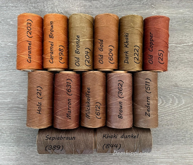 May include: A collection of fifteen cylindrical spools of thread in various shades of brown, tan, and copper. Each spool is labeled with a color name and a numerical code, including Caramel (203), Caramel Brown (498), Old Bronze (204), Old Gold (604), Dark Khaki (222), Old Copper (25), Holz (21), Marron (637), Milchkaffee (612), Brown (362), Zedern (511), Sepiabraun (389), and Khaki dunkel (844).
