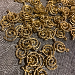 Può includere: Una collezione di ciondoli a spirale color oro, sparsi su una superficie di legno. Ogni ciondolo presenta un design a vortice strutturato con un piccolo anello nella parte superiore per il fissaggio. Probabilmente per la creazione di gioielli o altri progetti artigianali.