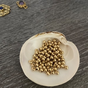 Könnte beinhalten: Eine Ansammlung kleiner, runder, goldfarbener Metallperlen, die in einer weißen Muschelschale liegen. Die Perlen haben eine glatte, reflektierende Oberfläche und ein mittiges Loch und werden häufig für Schmuckherstellung und Bastelprojekte verwendet.