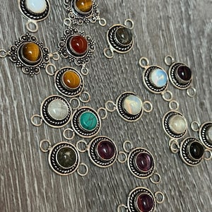 Può includere: Un assortimento di ciondoli con pietre preziose con montature in metallo color argento. I ciondoli presentano varie pietre colorate, tra cui occhio di tigre, turchese e ametista. I ciondoli sono disposti su una superficie di legno.