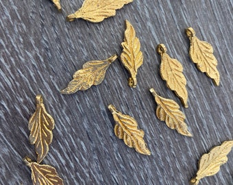 Könnte beinhalten: Auf einer Holzoberfläche liegen verstreut zahlreiche kleine, goldfarbene Blatt-Anhänger. Jeder Anhänger hat eine detaillierte, strukturierte Oberfläche, die einem echten Blatt ähnelt. Die Anhänger sind mit einer Schlaufe versehen, ideal für Schmuckherstellung oder Bastelprojekte.