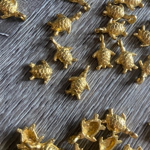 Op de afbeelding: Een verzameling kleine, goudkleurige schildpadbedels verspreid over een houten oppervlak. De bedels zijn gedetailleerd met getextureerde schilden en uitgestrekte ledematen, wat wijst op een sieraden- of knutselthema.