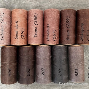 May include: Eleven cylindrical spools of thread arranged in two rows, featuring various shades of brown, tan, and taupe. Each spool is labeled with a color name and a numerical code, including Eisbraun 213, Sand dark 214, Taupe 366, Haselnuss 567, Braun 259, Mahogany 515, 496, 555, 207, 210, and 583.