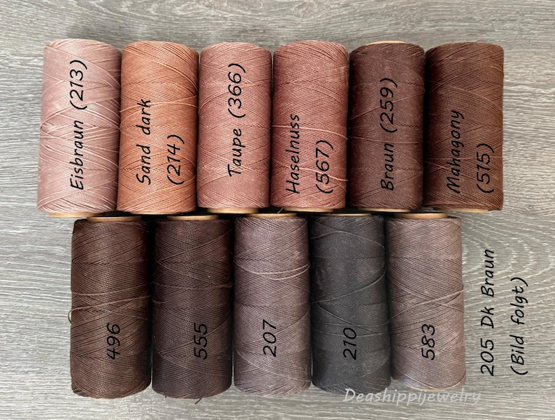 May include: Eleven cylindrical spools of thread arranged in two rows, featuring various shades of brown, tan, and taupe. Each spool is labeled with a color name and a numerical code, including Eisbraun 213, Sand dark 214, Taupe 366, Haselnuss 567, Braun 259, Mahogany 515, 496, 555, 207, 210, and 583.