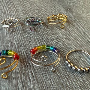 Könnte beinhalten: Sechs verschiedene Drahtringe mit bunten Perlen. Die Ringe sind aus Silber- und Golddraht gefertigt und haben unterschiedliche Perlmuster. Einige Ringe haben regenbogenfarbene Perlen, während andere silberne oder goldene Perlen haben.