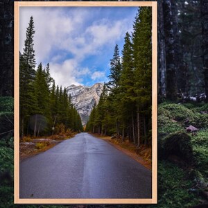 Pode incluir: Uma impressão emoldurada de uma paisagem com uma estrada que leva a uma cordilheira, ladeada por altas árvores perenes. O céu é azul com nuvens dispersas. A moldura é de cor castanho claro.