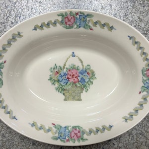 May include: White oval serving bowl with a floral design. The design features a basket of flowers with pink, blue, and yellow flowers. The bowl is trimmed with a yellow and green ribbon design.
