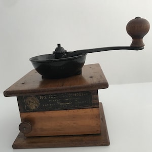 Può includere: Macinacaffè antico in legno con un meccanismo in ghisa nera e una maniglia in legno. Il macinacaffè ha un'etichetta che recita "No. 102 J.F. Woodill Nickel-Plated Coffee Mill Tap-Out Granulated Coffee Co.  Pat. Nov. 12, '95".