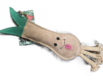 May include: A tan leather dog toy shaped like a long, thin vegetable with a green leaf on top. The toy has a smiling face with black eyes and a pink tongue sticking out. The toy has a rope tail with three strands.