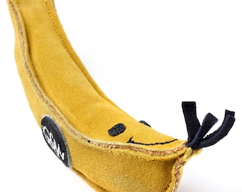 May include: A yellow suede banana-shaped dog toy with a black stitched smile and eyes. The toy has a black rope tail and a brown leather patch with the word "Grrr" on it.