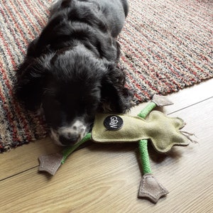 May include: A black and white puppy lying on a wooden floor next to a green and tan dog toy. The toy has a circular black patch with white text and a green rope body. The puppy is resting on a patterned rug.