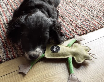May include: A black and white puppy lying on a wooden floor with a green and tan dog toy. The toy has a circular black patch with white text and a green rope body. The puppy is resting on a patterned rug.