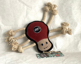May include: A red and brown dog toy shaped like a mushroom with a rope attached to each side. The toy has a black embroidered face and the text "GSM" on the top. The toy is on a white surface with a green and white tag that says "ECO Pet Toys".