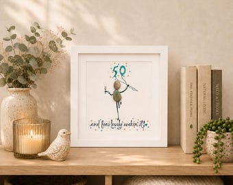 Puede incluir: Una pieza de arte mural cuadrada en marco blanco con una figura de piedras que sostiene una copa de champán y globos con el número 50. El texto dice 50 and fearlessly rockin' it! El diseño utiliza piedras lisas y líneas de tinta.