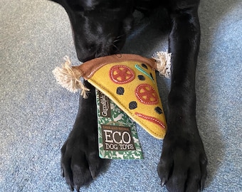 Puede incluir: Un perro negro jugando con un juguete para perros con forma de pizza amarillo y marrón. El juguete tiene ingredientes rojos y verdes y el texto "ECO DOG TOYS" está en el empaque.