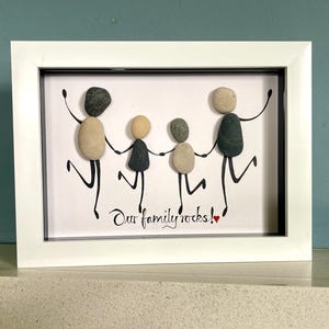 Op de afbeelding: Een ingelijst kunstwerk met een gezin van vier personen, afgebeeld met gladde, afgeronde stenen. De figuren houden elkaars handen vast en dansen, met zwarte lijnen voor armen en benen. De zin "Our family rocks!" staat eronder in cursief.