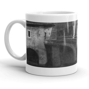 May include: A white ceramic mug featuring a black and white image of a stone building with an arched doorway and a window. The building is partially obscured by a tree.