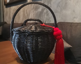 May include: Black woven wicker handbag with a red tassel. The bag has a round shape and a top handle.