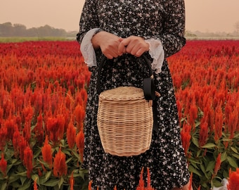 May include: A woman wearing a black floral dress with white lace trim holds a woven wicker basket with a black ribbon handle. The basket is in front of a field of red flowers.