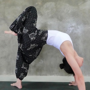 Peut inclure: Une personne portant un pantalon à motifs noir et blanc avec un débardeur blanc effectue une pose de yoga sur un tapis noir. La personne se penche en arrière, les jambes en l'air.